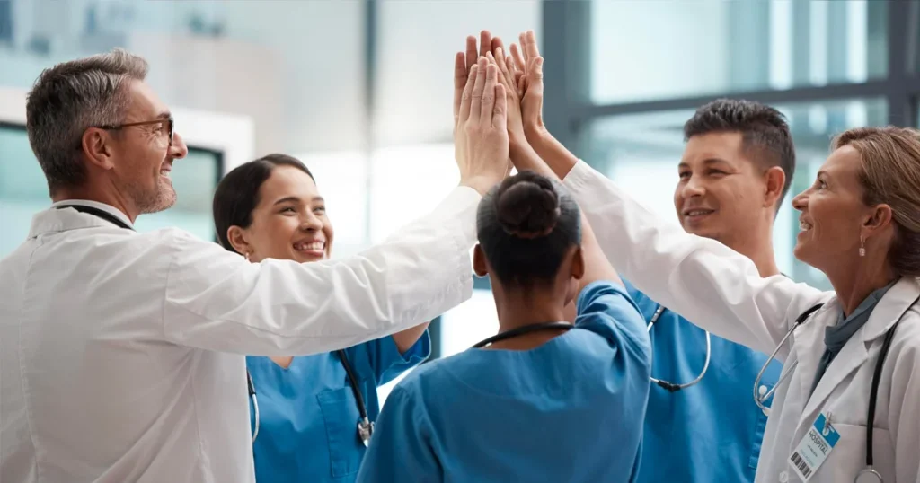 A diverse group of healthcare professionals, wearing scrubs and lab coats, celebrates with a high-five.